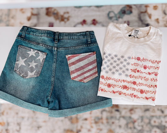 Cream-colored unisex t-shirt featuring a subtle and stylish American flag design made entirely of florals. The star field is composed of blue star-shaped flowers, while the red stripes are formed from intricate red floral garlands. This soft, vintage-inspired tee blends patriotic pride with a feminine, boho twist—perfect for summer celebrations like the Fourth of July, concerts, or casual weekends.