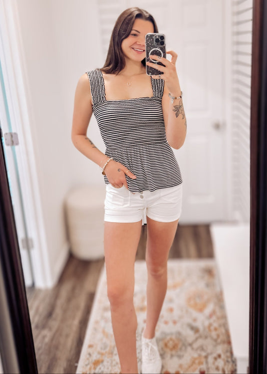 Woman wearing a black and white striped smocked peplum tank top with wide ruffled straps and a square neckline. The fitted smocked bodice creates a flattering silhouette, flaring into a flowy hem. Paired with light beige distressed denim shorts featuring frayed edges, this look is playful and breezy—perfect for sunny days, beach outings, or casual summer hangouts.