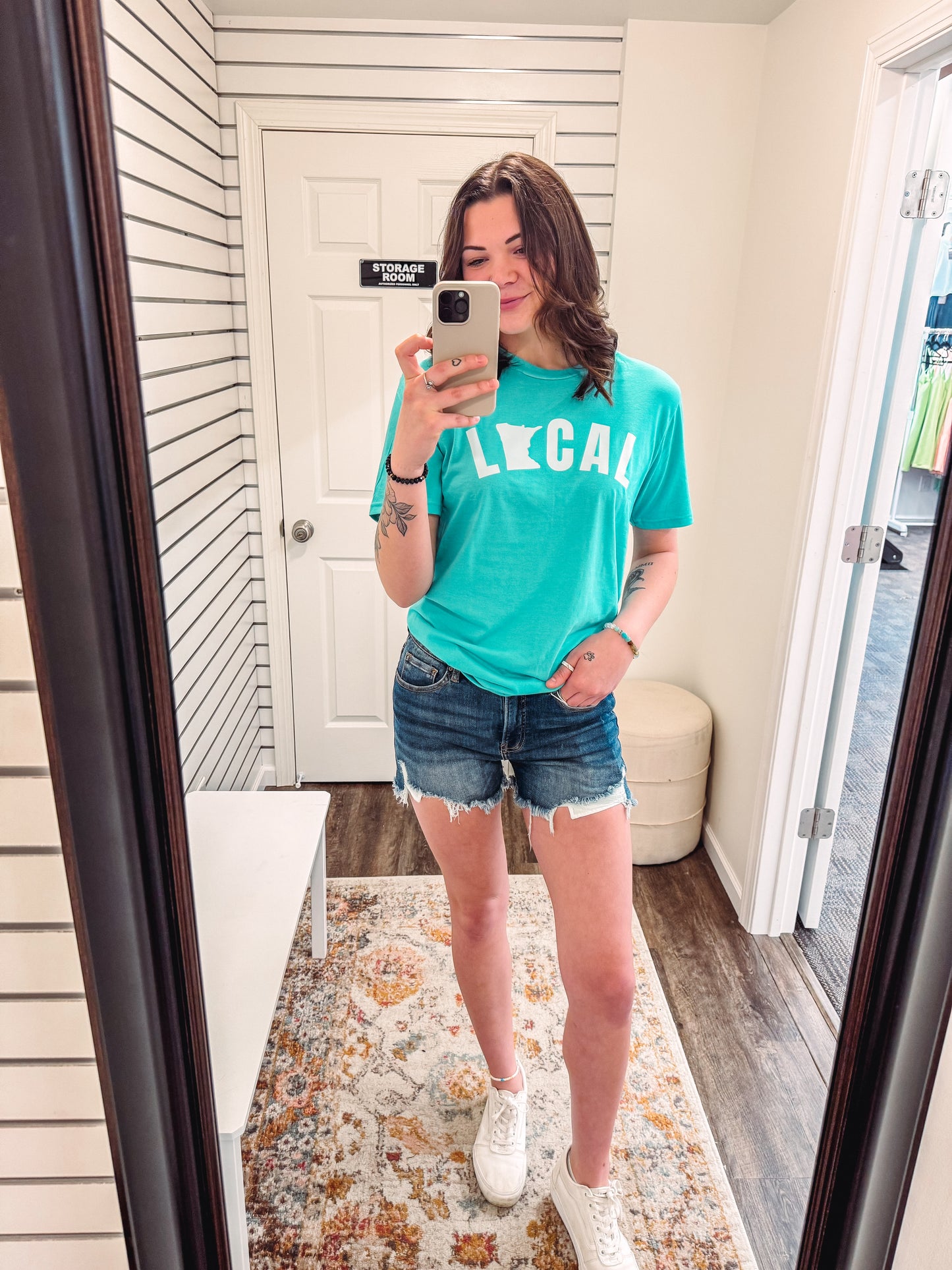 Woman wearing a turquoise crew neck graphic tee with the word “LOCAL” printed across the chest in bold white letters, featuring the silhouette of Minnesota in place of the “O.” The shirt has a relaxed fit and is styled with frayed denim shorts and white sneakers for a casual, small-town-proud summer look. Perfect for repping your roots, weekend events, or supporting your favorite local spots with comfy flair.
