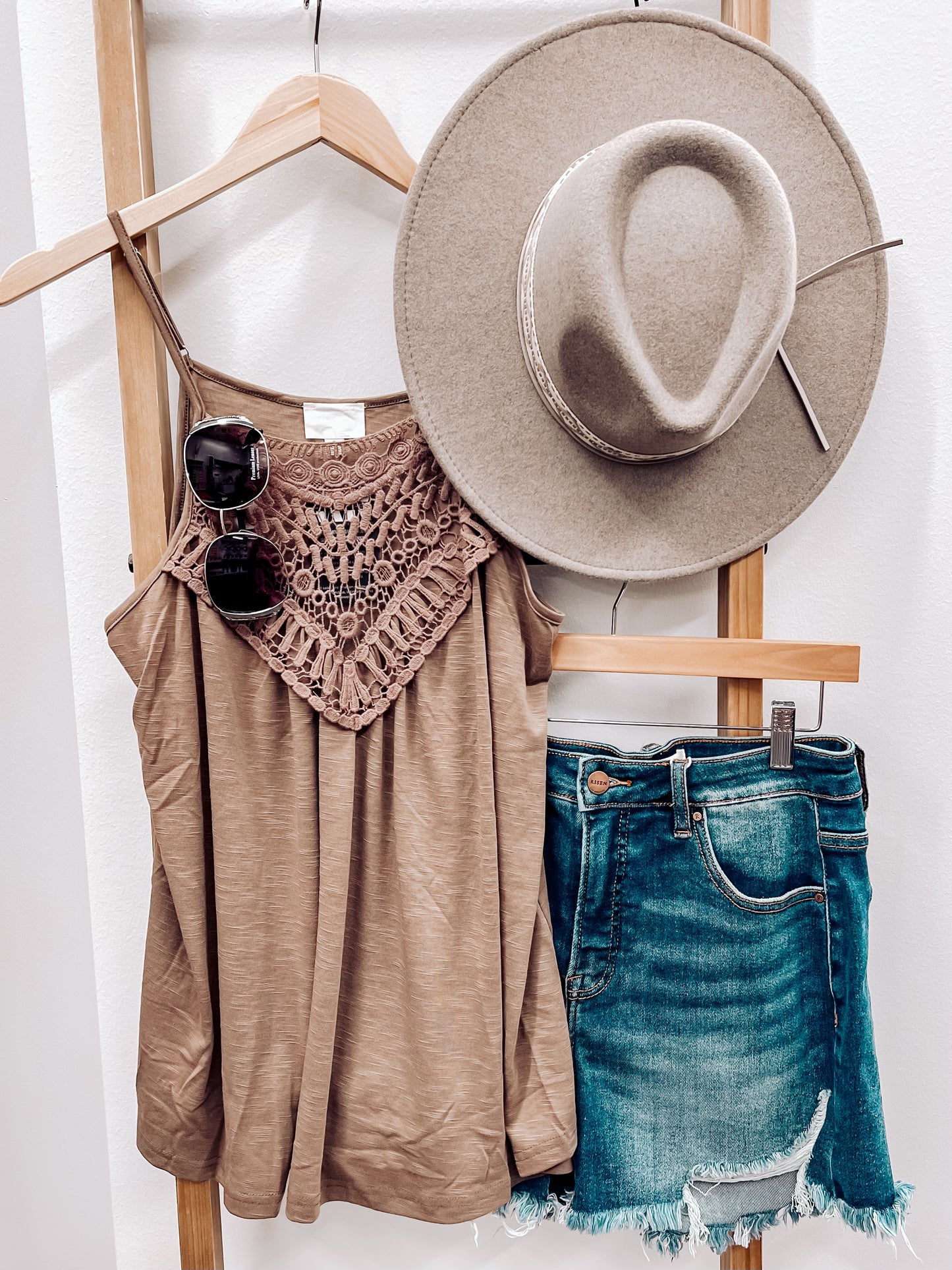 Woman taking a mirror selfie wearing a mocha brown halter tank top with intricate crochet lace detailing across the neckline and chest. The lightweight, flowy fabric drapes softly with a relaxed fit. Thin spaghetti straps and subtle pleating add to its breezy, feminine vibe. She pairs it with high-rise, frayed hem denim shorts—perfect for a casual yet stylish summer outfit.