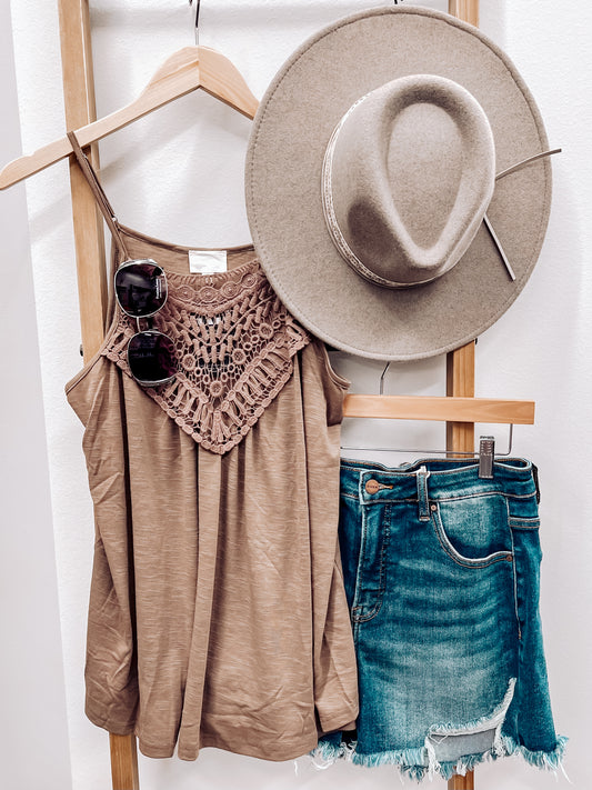 Woman taking a mirror selfie wearing a mocha brown halter tank top with intricate crochet lace detailing across the neckline and chest. The lightweight, flowy fabric drapes softly with a relaxed fit. Thin spaghetti straps and subtle pleating add to its breezy, feminine vibe. She pairs it with high-rise, frayed hem denim shorts—perfect for a casual yet stylish summer outfit.