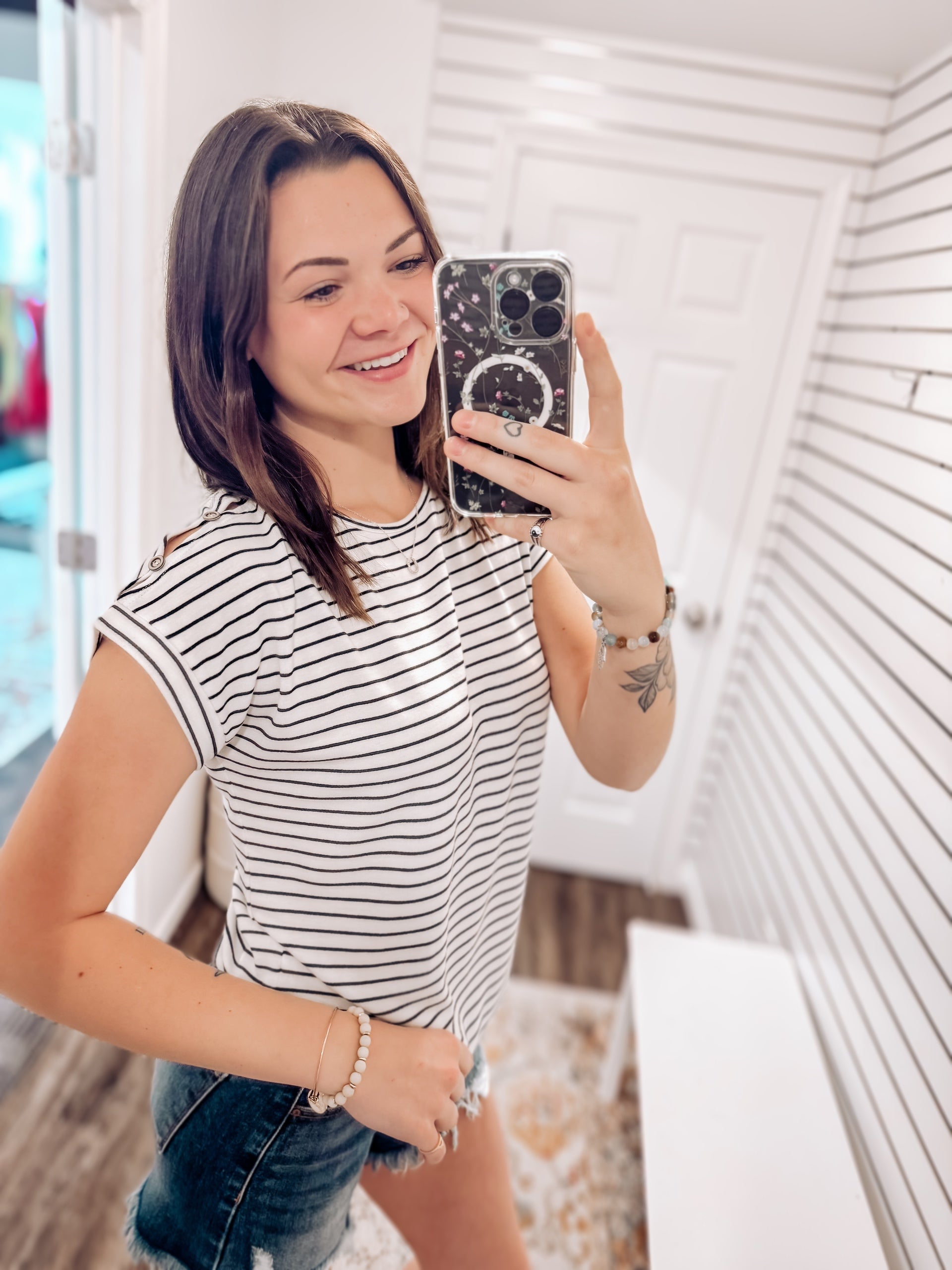 Woman wearing a white short-sleeve top with thin black horizontal stripes and rolled cuffs. The shirt features a relaxed, flowy fit with a crew neckline and button detail on the sleeves. Styled with white cuffed shorts, this effortless everyday tee is perfect for warm weather, offering a classic nautical-inspired look that’s both casual and polished.