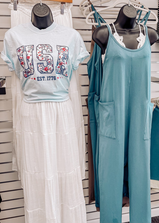 Light blue unisex t-shirt featuring bold "USA" block letters filled with a red, white, and blue floral pattern for a faux-embroidered look. Below, "EST. 1776" is printed in navy with red stars, celebrating American heritage. Classic crew neck and short sleeves offer a comfortable fit, perfect for patriotic holidays like the Fourth of July or Memorial Day.