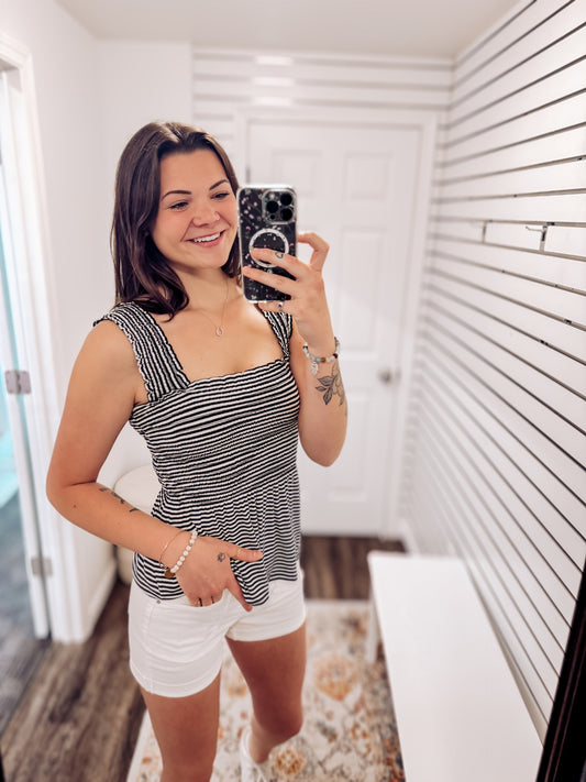Woman wearing a black and white striped smocked peplum tank top with wide ruffled straps and a square neckline. The fitted smocked bodice creates a flattering silhouette, flaring into a flowy hem. Paired with light beige distressed denim shorts featuring frayed edges, this look is playful and breezy—perfect for sunny days, beach outings, or casual summer hangouts.