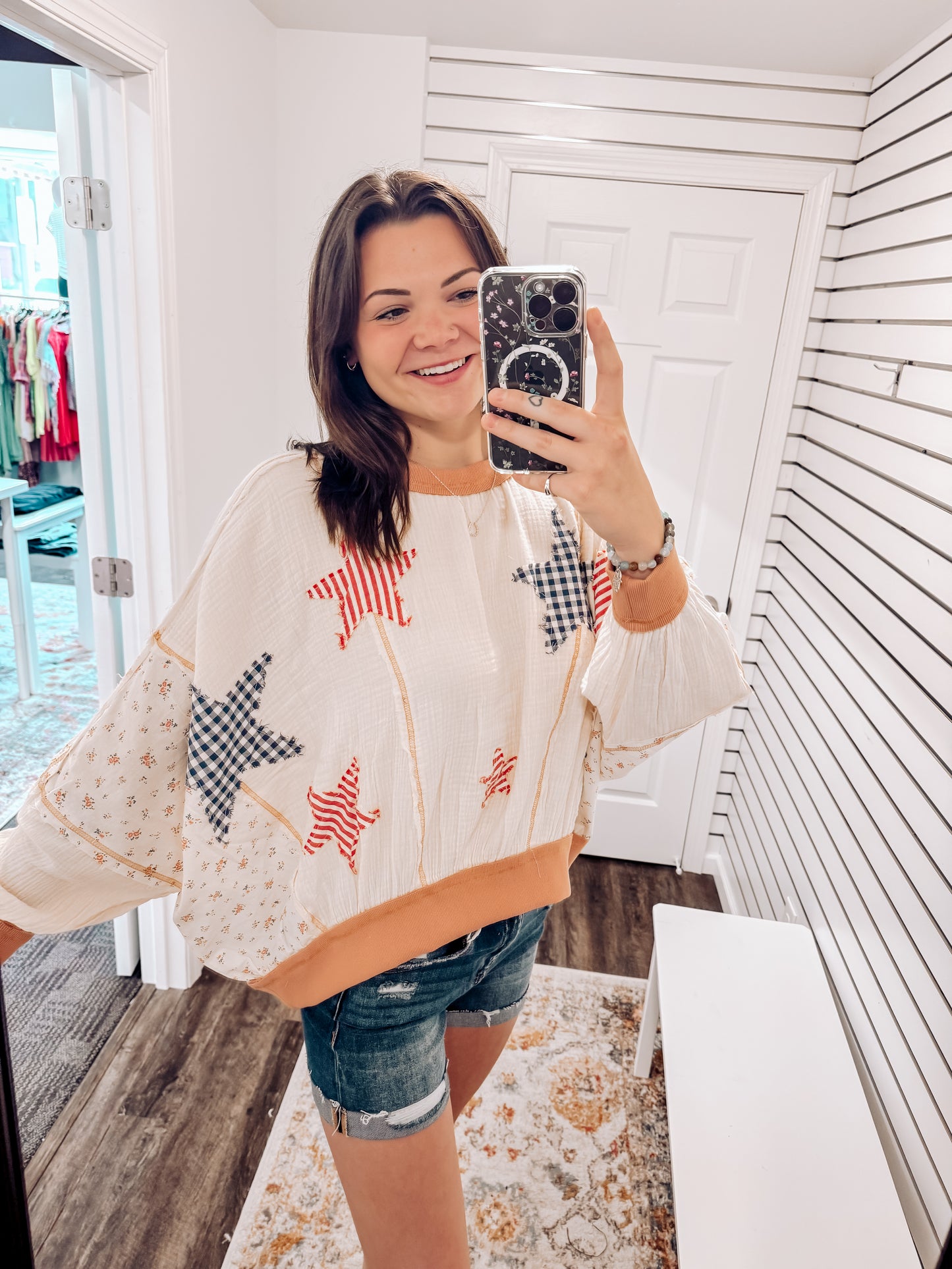 Woman wearing a cream textured pullover with raw seam detailing and patchwork star appliqués in red stripes and blue gingham. Features blush pink ribbed cuffs and hem, floral-print sleeves, and a relaxed, oversized fit. Styled with distressed light-wash denim shorts and a wide-brim cream hat, this cozy top delivers vintage Americana vibes with a soft, boho twist—perfect for patriotic holidays or laid-back summer days.