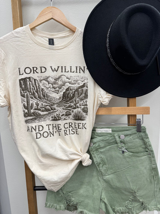 Cream-colored unisex graphic tee featuring a vintage-style desert landscape with cacti, mesas, and cloud-filled skies. The bold black text reads “Lord Willin’ And The Creek Don’t Rise,” giving major western vibes with a faith-filled twist. Styled here with sage green frayed shorts and a black wide-brim hat for an effortlessly cool, rustic look. Perfect for rodeo nights, Sunday coffee runs, or dusty backroad adventures.