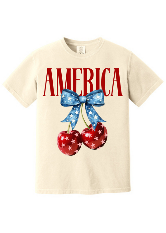 Cream-colored unisex t-shirt featuring bold red “AMERICA” text across the chest. Below, two glossy red cherries with white stars hang from a vibrant blue bow decorated with a star pattern, creating a playful and patriotic summer design. Classic crew neck and short sleeves make it perfect for 4th of July festivities or showing off your American pride with a sweet twist.