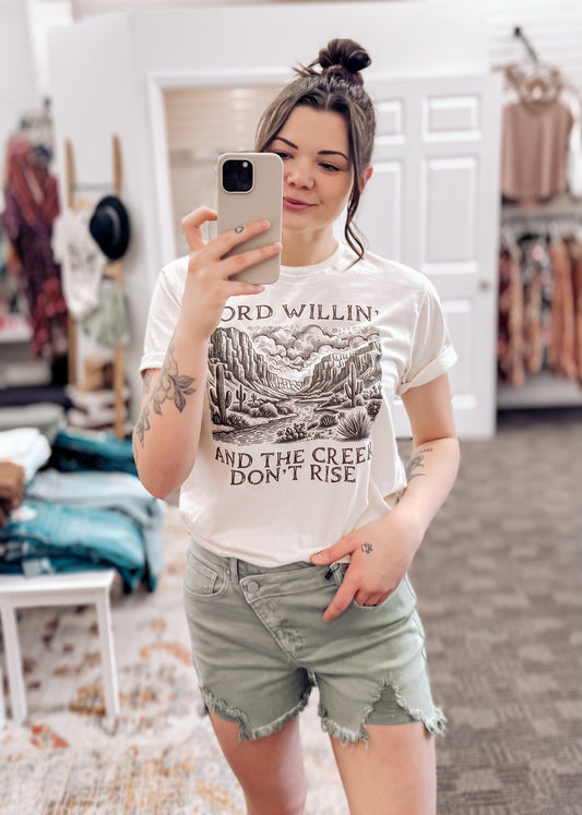 Cream-colored unisex graphic tee featuring a vintage-style desert landscape with cacti, mesas, and cloud-filled skies. The bold black text reads “Lord Willin’ And The Creek Don’t Rise,” giving major western vibes with a faith-filled twist. Styled here with sage green frayed shorts and a black wide-brim hat for an effortlessly cool, rustic look. Perfect for rodeo nights, Sunday coffee runs, or dusty backroad adventures.