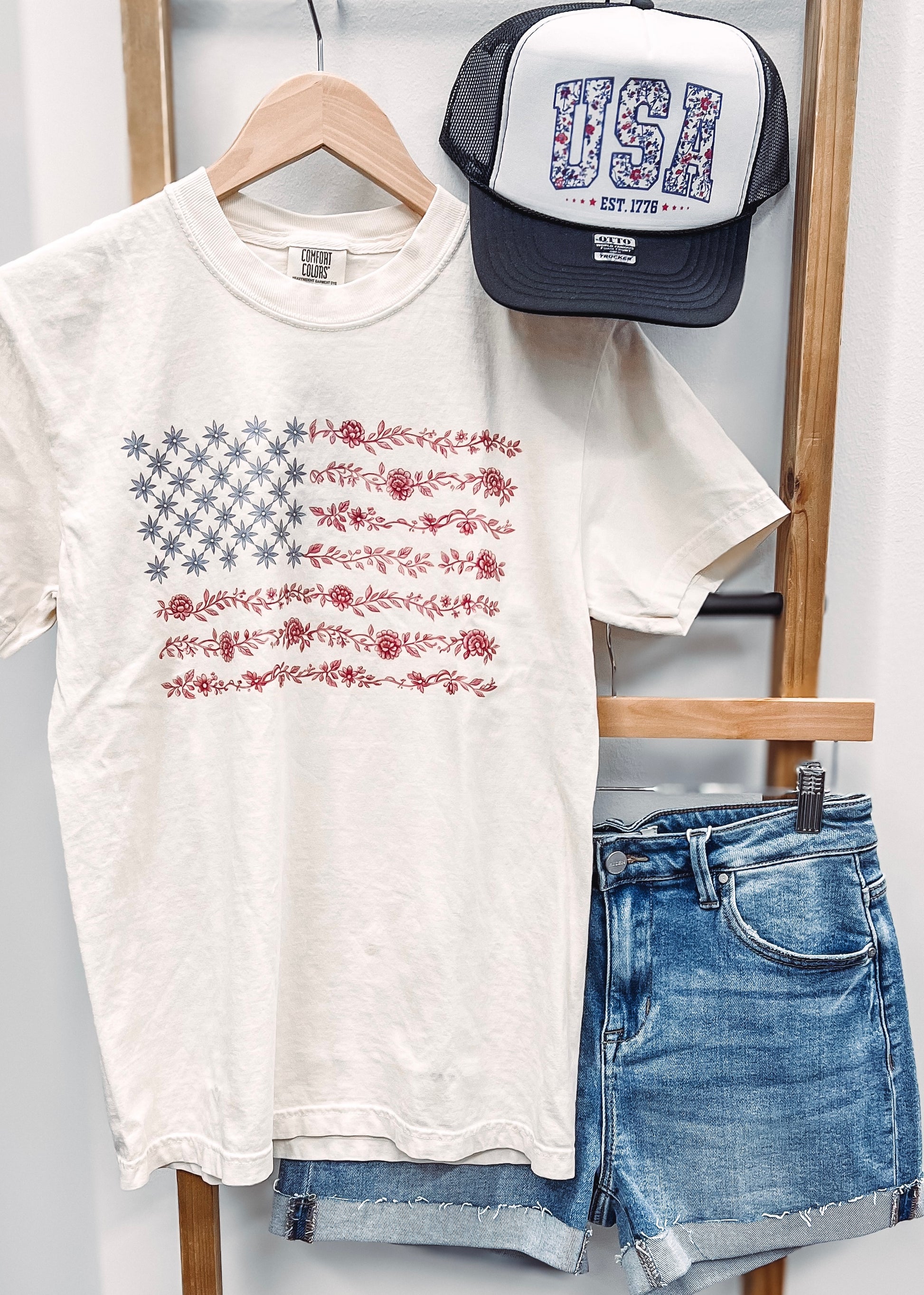 Cream-colored unisex t-shirt featuring a subtle and stylish American flag design made entirely of florals. The star field is composed of blue star-shaped flowers, while the red stripes are formed from intricate red floral garlands. This soft, vintage-inspired tee blends patriotic pride with a feminine, boho twist—perfect for summer celebrations like the Fourth of July, concerts, or casual weekends.