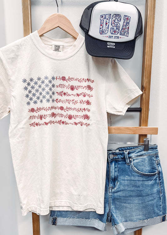 Cream-colored unisex t-shirt featuring a subtle and stylish American flag design made entirely of florals. The star field is composed of blue star-shaped flowers, while the red stripes are formed from intricate red floral garlands. This soft, vintage-inspired tee blends patriotic pride with a feminine, boho twist—perfect for summer celebrations like the Fourth of July, concerts, or casual weekends.