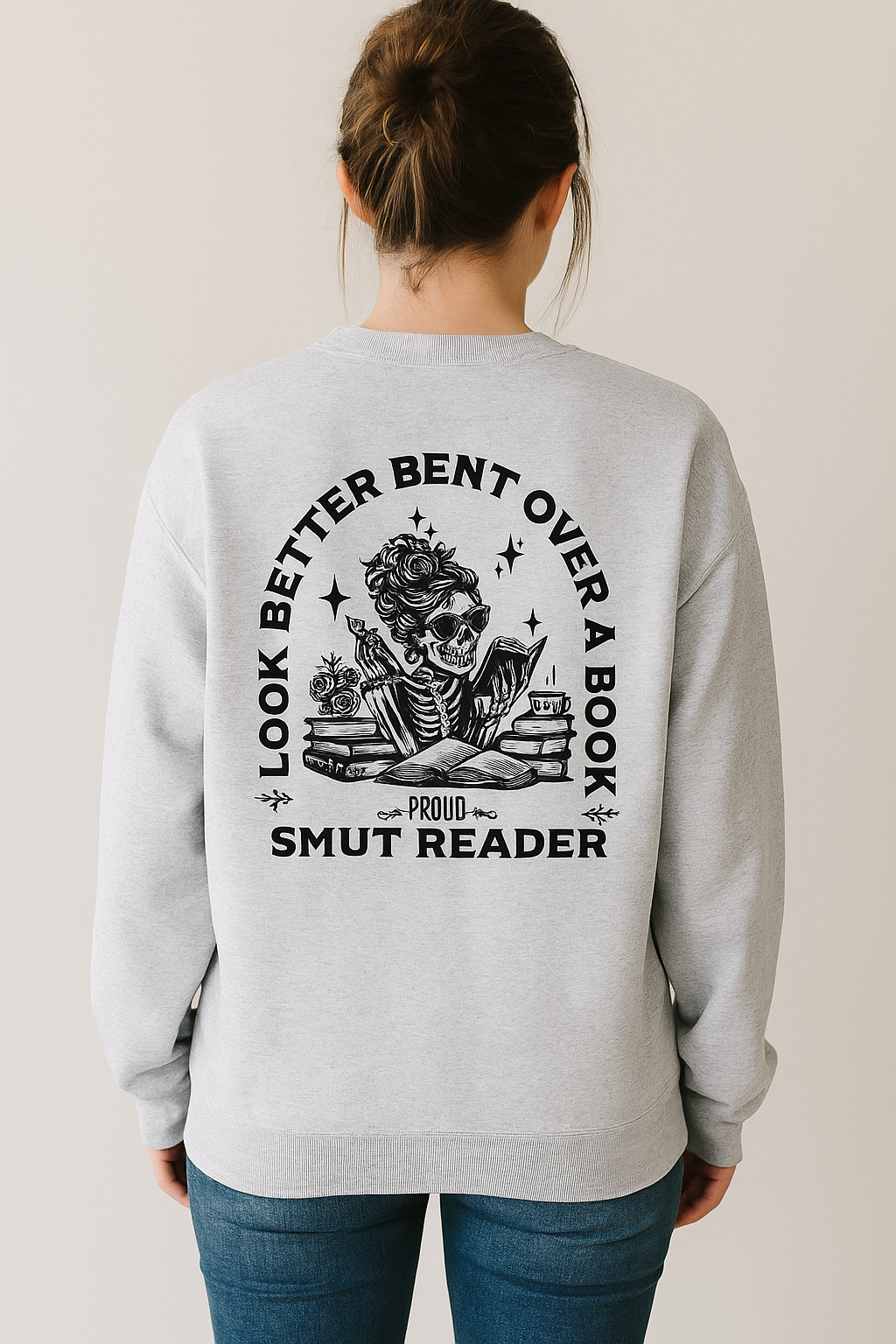 Light gray crewneck sweatshirt featuring bold black bookish graphics. The front chest has a small open-book design with a hand and the phrase “Spread Those Pages.” The back showcases a full skeleton girl reading, surrounded by stacked books, coffee, and sparkles. Large text reads: “I Look Better Bent Over a Book – Proud Smut Reader.” A cozy, cheeky must-have for spicy book lovers with a sense of humor.