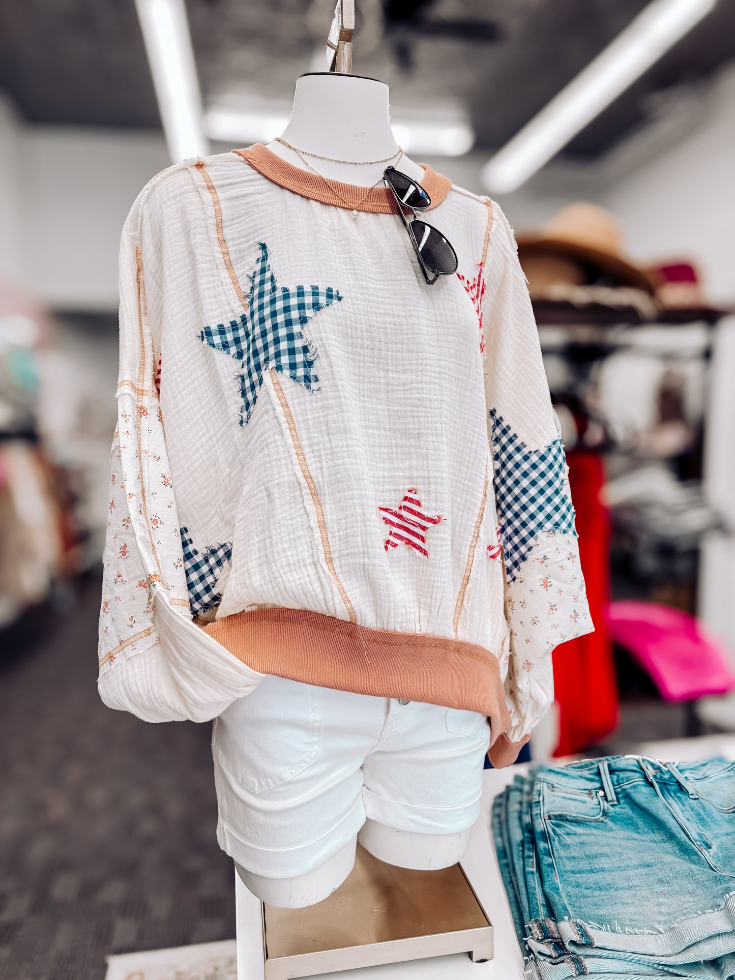 Woman wearing a cream textured pullover with raw seam detailing and patchwork star appliqués in red stripes and blue gingham. Features blush pink ribbed cuffs and hem, floral-print sleeves, and a relaxed, oversized fit. Styled with distressed light-wash denim shorts and a wide-brim cream hat, this cozy top delivers vintage Americana vibes with a soft, boho twist—perfect for patriotic holidays or laid-back summer days.