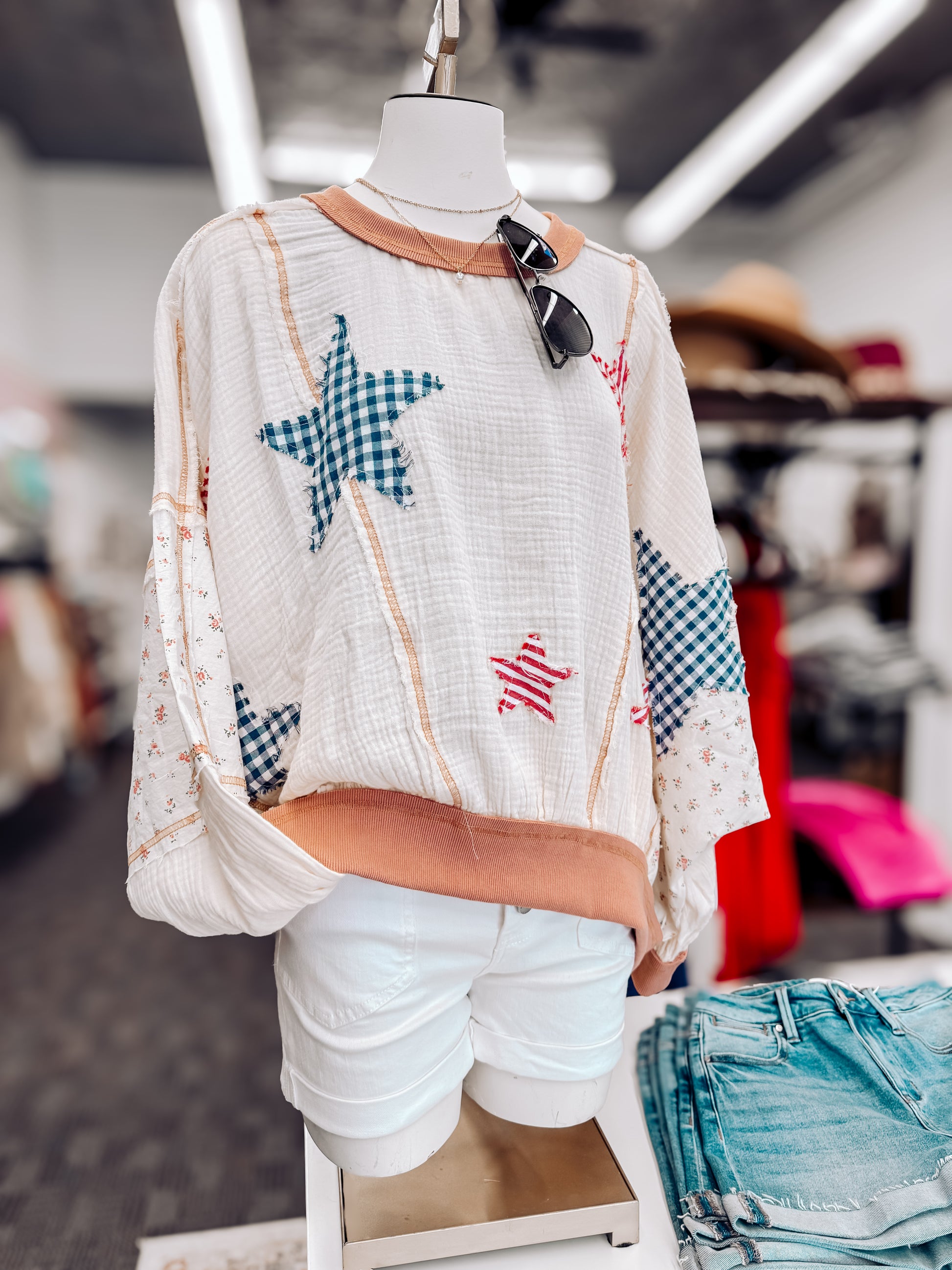 Woman wearing a cream textured pullover with raw seam detailing and patchwork star appliqués in red stripes and blue gingham. Features blush pink ribbed cuffs and hem, floral-print sleeves, and a relaxed, oversized fit. Styled with distressed light-wash denim shorts and a wide-brim cream hat, this cozy top delivers vintage Americana vibes with a soft, boho twist—perfect for patriotic holidays or laid-back summer days.