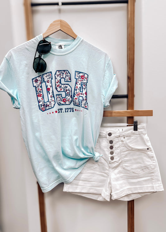 Light blue unisex t-shirt featuring bold "USA" block letters filled with a red, white, and blue floral pattern for a faux-embroidered look. Below, "EST. 1776" is printed in navy with red stars, celebrating American heritage. Classic crew neck and short sleeves offer a comfortable fit, perfect for patriotic holidays like the Fourth of July or Memorial Day.