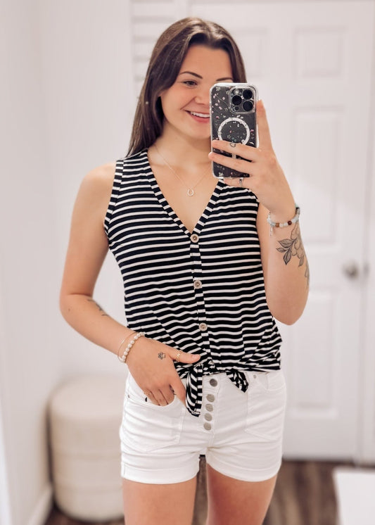 Woman wearing a sleeveless black and white horizontal striped tank top with a deep V-neckline and functional wooden buttons down the front. The top features a trendy front tie detail at the hem, creating a casual, flattering silhouette. Made from a soft, lightweight fabric, it's styled with medium-wash denim jeans—perfect for laid-back summer days, weekend outings, or everyday wear.