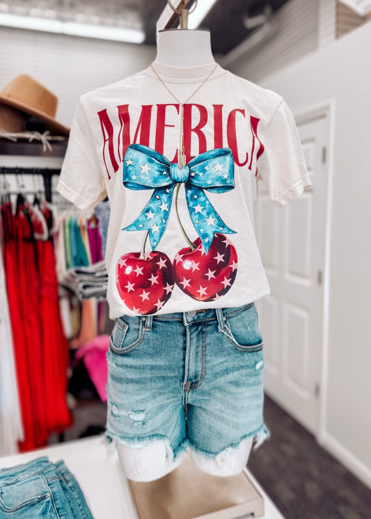 Cream-colored unisex t-shirt featuring bold red “AMERICA” text across the chest. Below, two glossy red cherries with white stars hang from a vibrant blue bow decorated with a star pattern, creating a playful and patriotic summer design. Classic crew neck and short sleeves make it perfect for 4th of July festivities or showing off your American pride with a sweet twist.