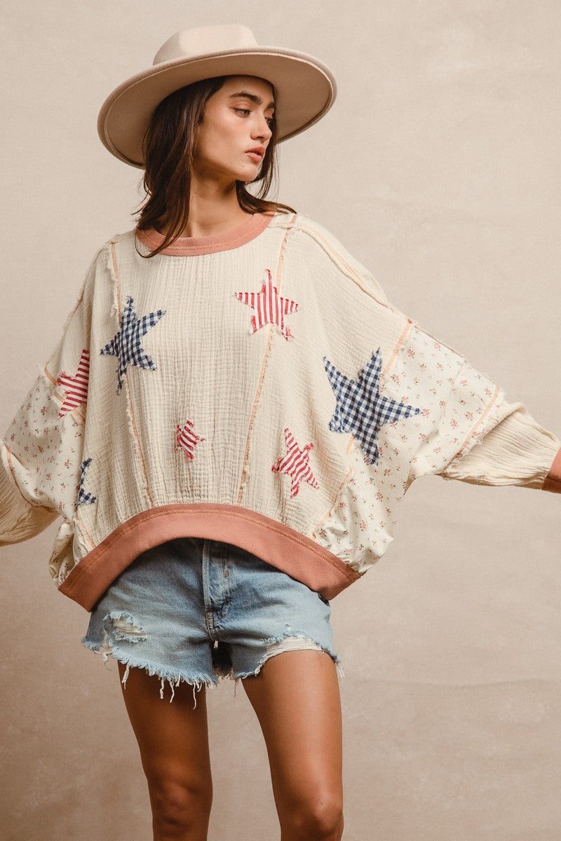 Woman wearing a cream textured pullover with raw seam detailing and patchwork star appliqués in red stripes and blue gingham. Features blush pink ribbed cuffs and hem, floral-print sleeves, and a relaxed, oversized fit. Styled with distressed light-wash denim shorts and a wide-brim cream hat, this cozy top delivers vintage Americana vibes with a soft, boho twist—perfect for patriotic holidays or laid-back summer days.