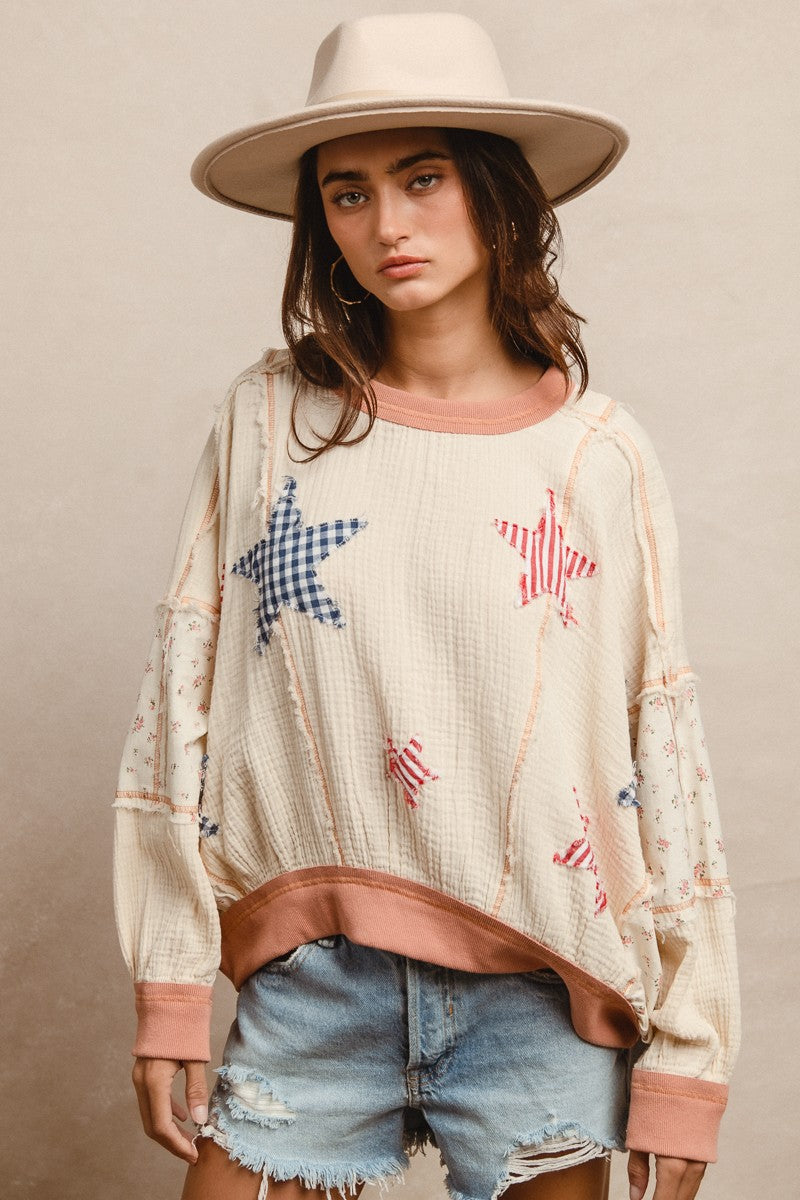 Woman wearing a cream textured pullover with raw seam detailing and patchwork star appliqués in red stripes and blue gingham. Features blush pink ribbed cuffs and hem, floral-print sleeves, and a relaxed, oversized fit. Styled with distressed light-wash denim shorts and a wide-brim cream hat, this cozy top delivers vintage Americana vibes with a soft, boho twist—perfect for patriotic holidays or laid-back summer days.