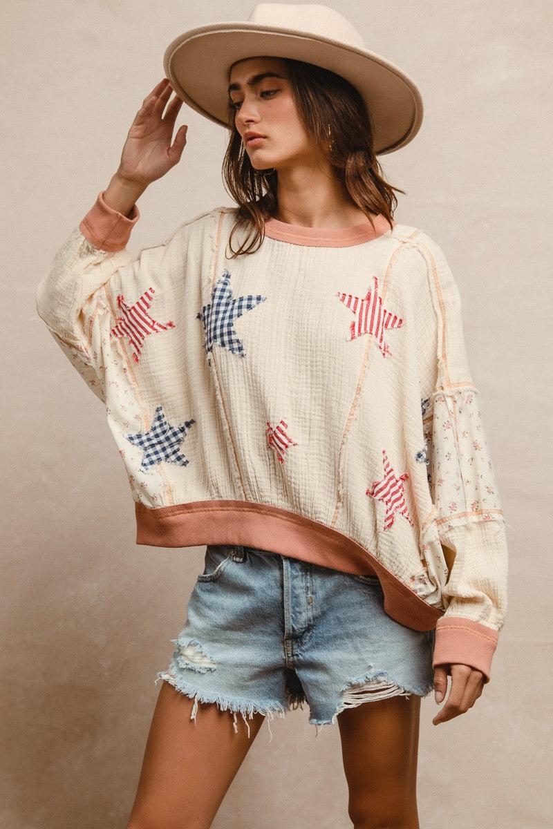 Woman wearing a cream textured pullover with raw seam detailing and patchwork star appliqués in red stripes and blue gingham. Features blush pink ribbed cuffs and hem, floral-print sleeves, and a relaxed, oversized fit. Styled with distressed light-wash denim shorts and a wide-brim cream hat, this cozy top delivers vintage Americana vibes with a soft, boho twist—perfect for patriotic holidays or laid-back summer days.