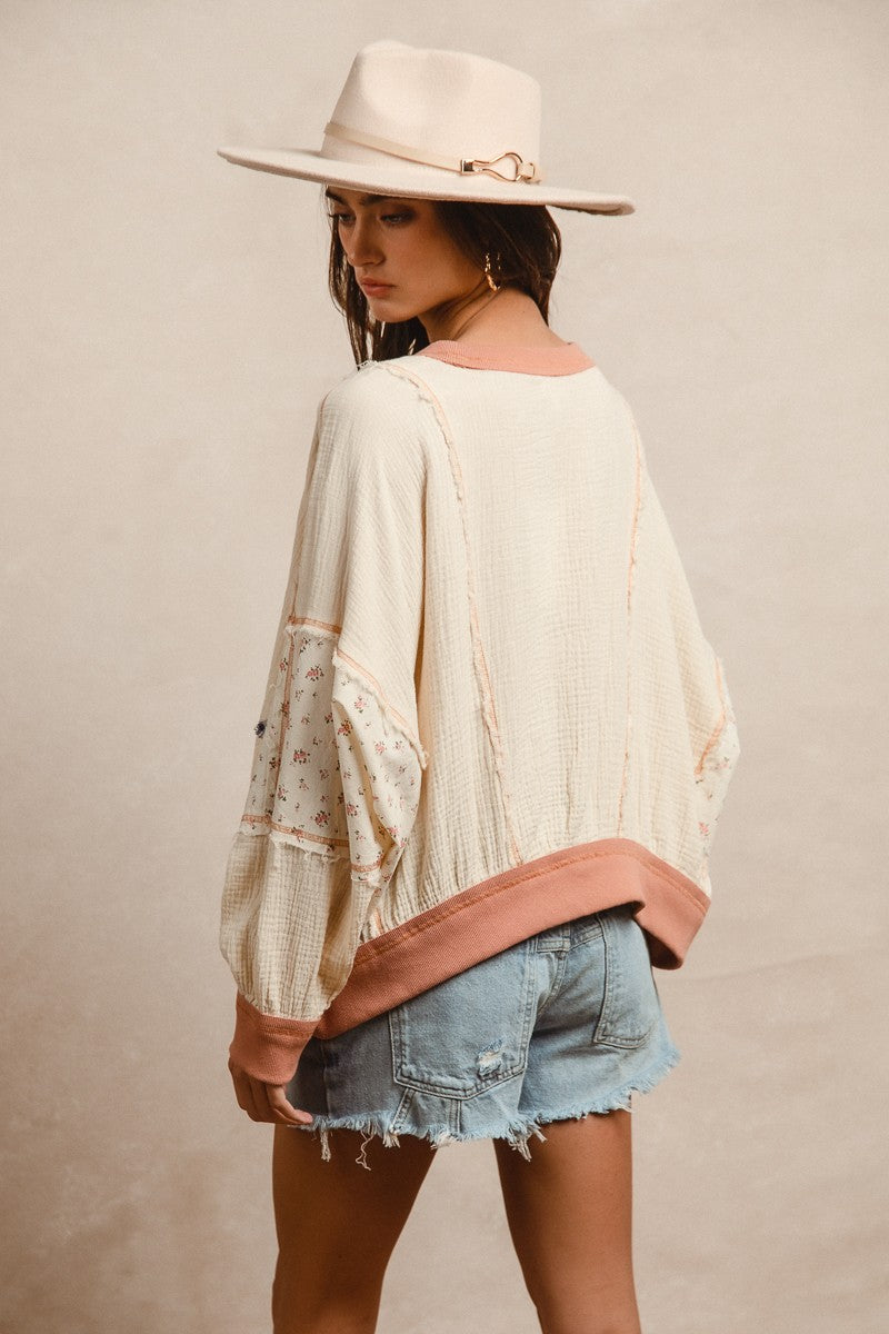 Woman wearing a cream textured pullover with raw seam detailing and patchwork star appliqués in red stripes and blue gingham. Features blush pink ribbed cuffs and hem, floral-print sleeves, and a relaxed, oversized fit. Styled with distressed light-wash denim shorts and a wide-brim cream hat, this cozy top delivers vintage Americana vibes with a soft, boho twist—perfect for patriotic holidays or laid-back summer days.