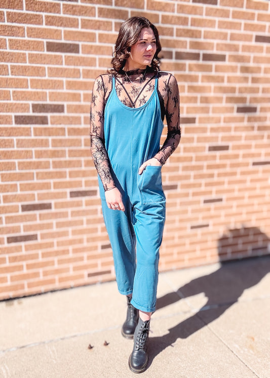 Dusty blue relaxed-fit jumpsuit with adjustable spaghetti straps and a scoop neckline. Features a slouchy, drop-crotch silhouette with deep side pockets and cuffed hems for a laid-back, comfy vibe. Made with ultra-soft stretch knit fabric, it’s perfect for lounging, errands, or layering over a tank or bralette for easy, everyday style.