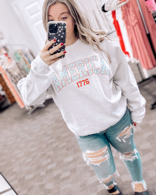 Light gray crewneck sweatshirt featuring bold varsity-style block lettering that reads "AMERICA" in sky blue with white stars, and “1776” in bright red beneath. Made with a soft fleece interior and relaxed fit for everyday comfort. Pairs perfectly with distressed denim for a casual patriotic vibe. Ideal for summer nights, Fourth of July, or anyone repping red, white, and blue with a cozy twist.