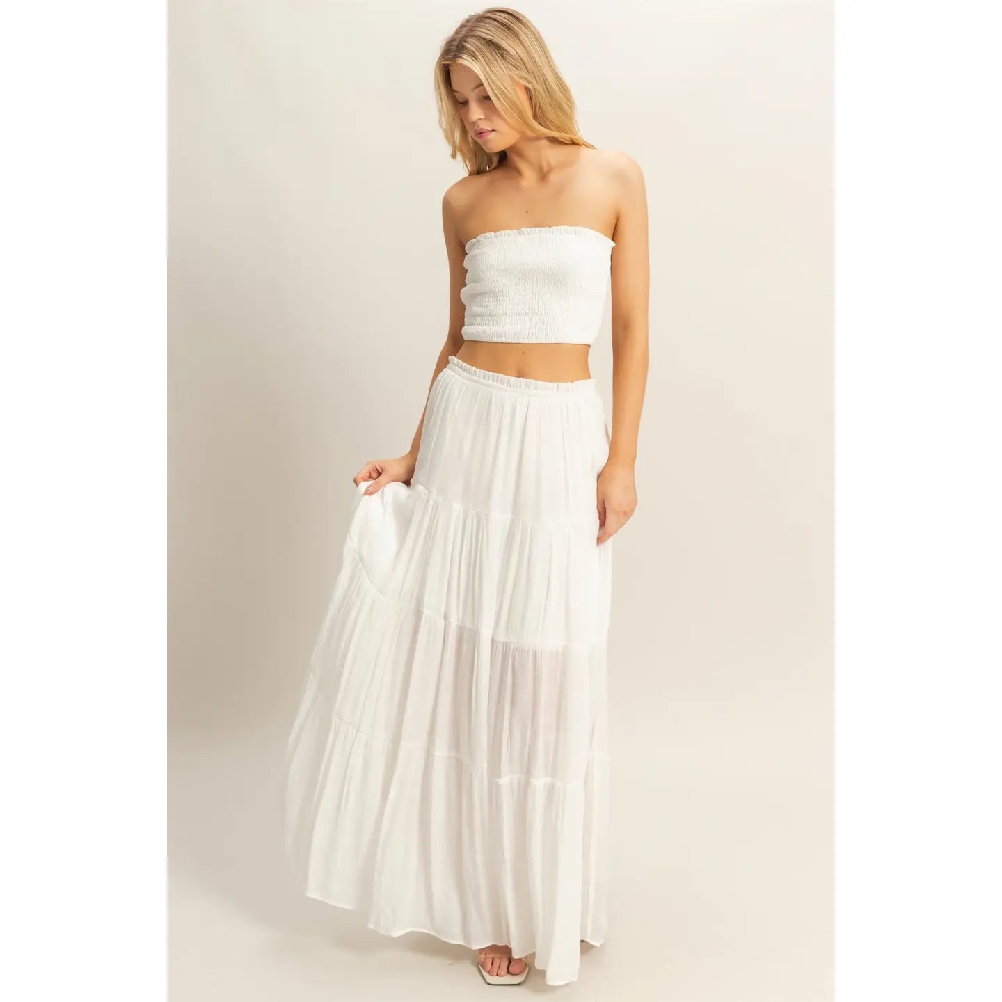 Woman wearing a white strapless smocked crop top and matching flowy tiered maxi skirt, standing against a neutral background.