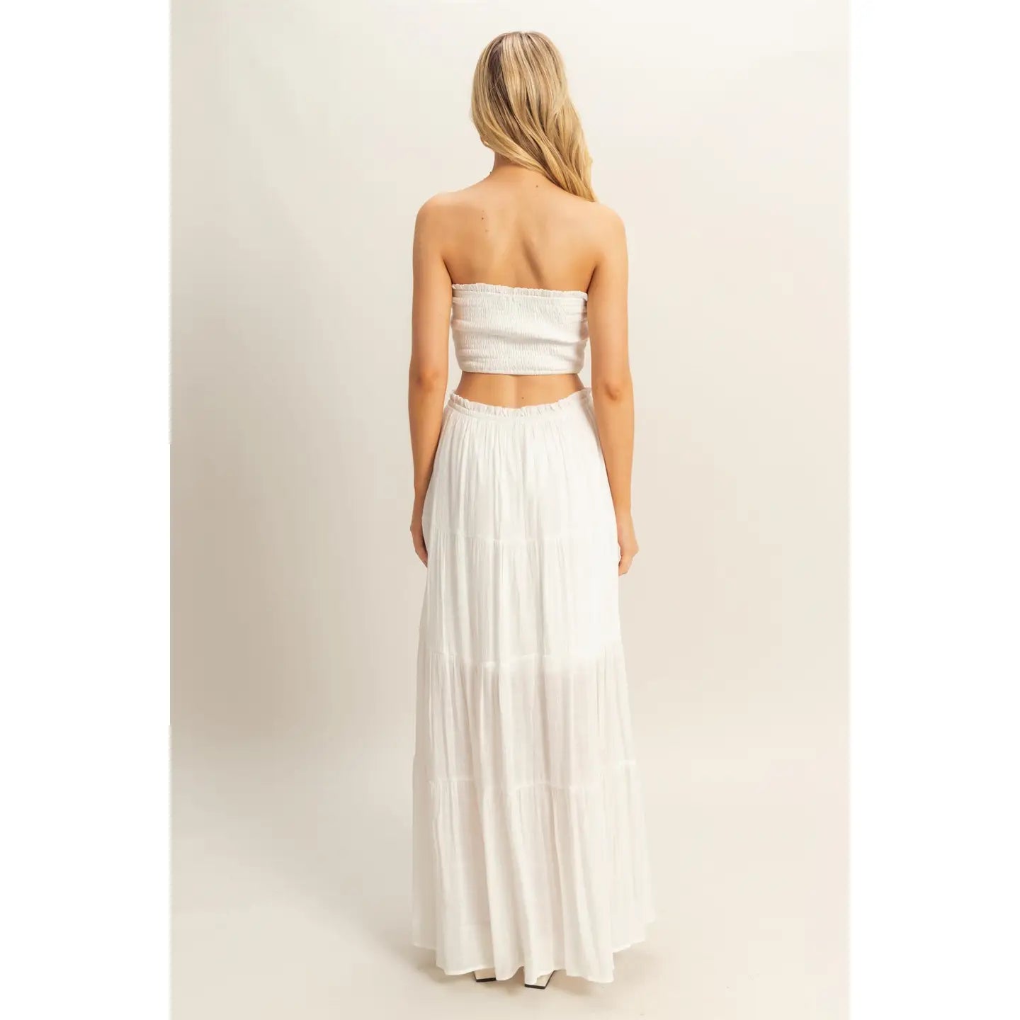 Woman wearing a white strapless smocked crop top and matching flowy tiered maxi skirt, standing against a neutral background.