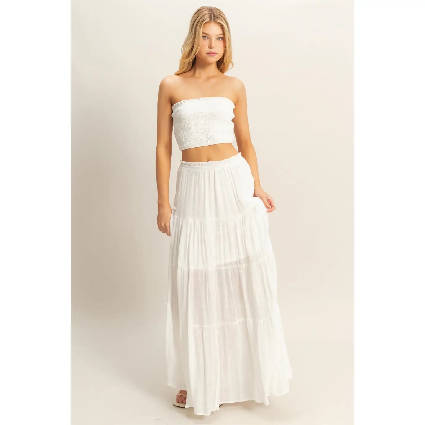 Woman wearing a white strapless smocked crop top and matching flowy tiered maxi skirt, standing against a neutral background.