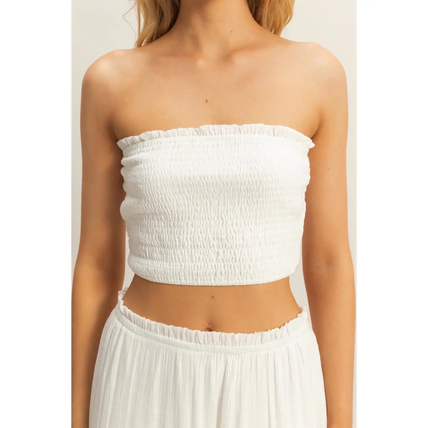 Woman wearing a white strapless smocked crop top and matching flowy tiered maxi skirt, standing against a neutral background.