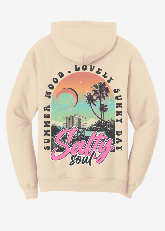 Cream-colored unisex hoodie featuring a large, colorful back graphic. The design includes a vibrant sunset sky in orange, teal, and lavender tones with a crescent moon, tall palm trees, a lifeguard tower, and ocean waves. The text "Summer Mood, Lovely Sunny Day" curves around the top of the image, while the bold phrase "Salty Soul" is prominently displayed at the bottom in large pink and white retro-style script. The graphic has a vintage, worn-in texture, creating a laid-back coastal aesthetic.