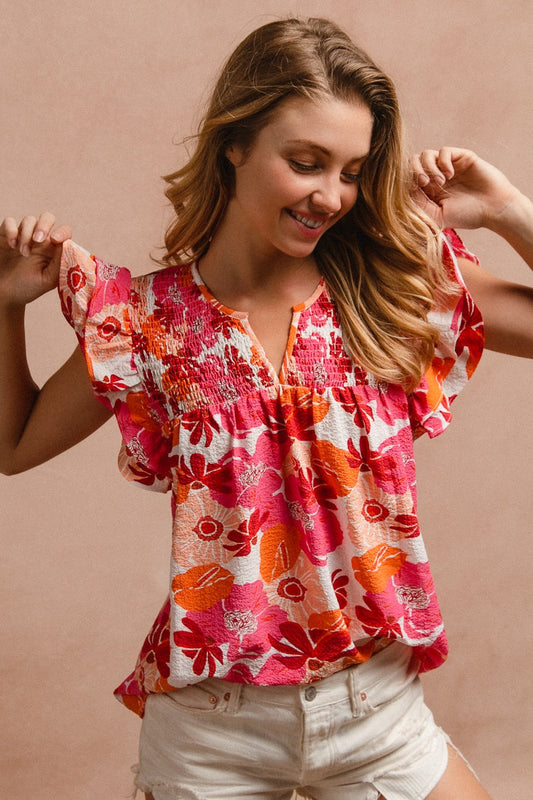 Woman wearing a vibrant floral short-sleeve top with a textured, crinkle fabric. The blouse features shades of pink, red, orange, and white in a bold floral print, with ruffled sleeves and a smocked detail across the chest. A split V-neckline adds a flattering touch, and the relaxed fit pairs effortlessly with her high-rise white distressed denim shorts. A bright, cheerful piece perfect for summer days, vacations, or casual outings.