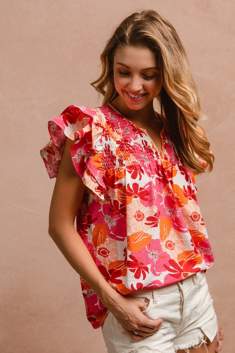 Woman wearing a vibrant floral short-sleeve top with a textured, crinkle fabric. The blouse features shades of pink, red, orange, and white in a bold floral print, with ruffled sleeves and a smocked detail across the chest. A split V-neckline adds a flattering touch, and the relaxed fit pairs effortlessly with her high-rise white distressed denim shorts. A bright, cheerful piece perfect for summer days, vacations, or casual outings.