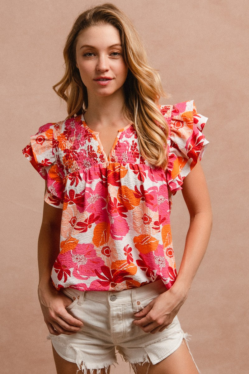 Woman wearing a vibrant floral short-sleeve top with a textured, crinkle fabric. The blouse features shades of pink, red, orange, and white in a bold floral print, with ruffled sleeves and a smocked detail across the chest. A split V-neckline adds a flattering touch, and the relaxed fit pairs effortlessly with her high-rise white distressed denim shorts. A bright, cheerful piece perfect for summer days, vacations, or casual outings.