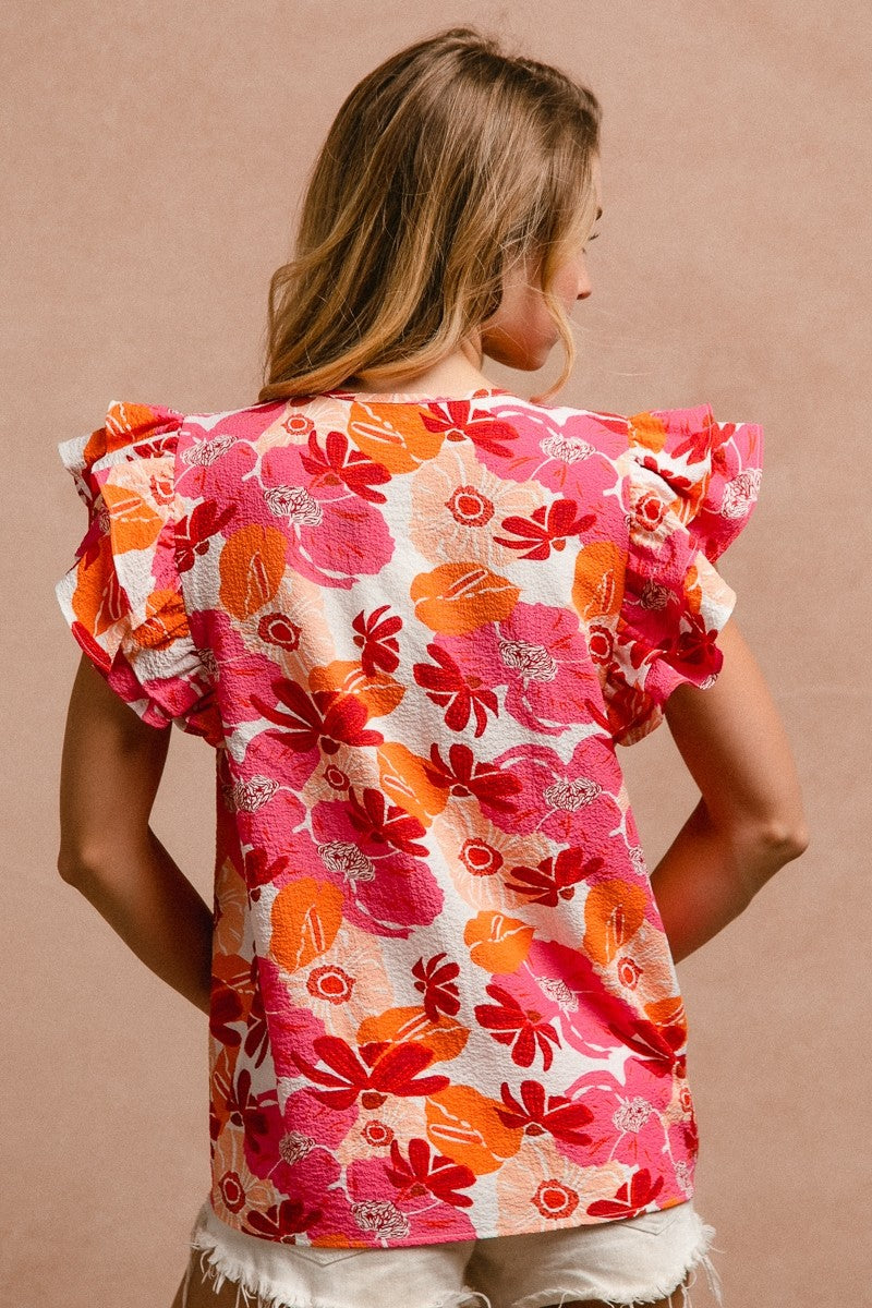 Woman wearing a vibrant floral short-sleeve top with a textured, crinkle fabric. The blouse features shades of pink, red, orange, and white in a bold floral print, with ruffled sleeves and a smocked detail across the chest. A split V-neckline adds a flattering touch, and the relaxed fit pairs effortlessly with her high-rise white distressed denim shorts. A bright, cheerful piece perfect for summer days, vacations, or casual outings.