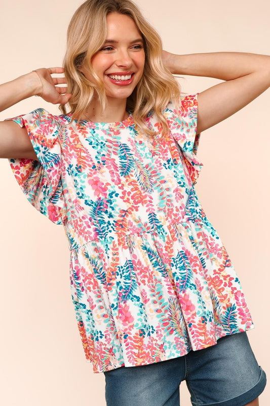 Woman wearing a vibrant, multicolored floral print babydoll top with a round neckline and flutter sleeves. The top features a loose, flowy silhouette with a gathered waist seam that adds volume and movement. The pattern includes shades of coral, teal, pink, orange, and blue against a white background, creating a playful, summery look. She pairs it with medium-wash denim shorts. Her long blonde hair is styled in soft waves, and she stands against a light peach backdrop that complements the warm tones of the