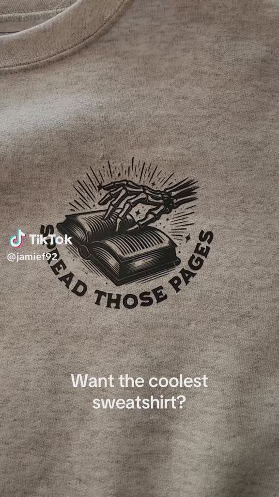 Light gray crewneck sweatshirt featuring bold black bookish graphics. The front chest has a small open-book design with a hand and the phrase “Spread Those Pages.” The back showcases a full skeleton girl reading, surrounded by stacked books, coffee, and sparkles. Large text reads: “I Look Better Bent Over a Book – Proud Smut Reader.” A cozy, cheeky must-have for spicy book lovers with a sense of humor.
