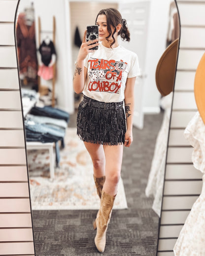 Beige graphic tee featuring a bold red "Tears of a Cowboy" slogan layered over a black-and-white rodeo newspaper print. Accents include dice, a horseshoe, and distressed lettering for an edgy Western-meets-streetwear vibe. Perfect for rodeo rebels, heartbreak cowgirls, or anyone living on the wild side of the saddle.