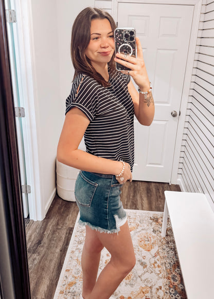 Woman wearing a black short-sleeve top with thin white horizontal stripes, a classic crew neckline, and rolled cuff sleeves with button accents. The top features a relaxed, drapey fit that pairs effortlessly with white cuffed shorts for a crisp, casual look. Perfect for everyday wear, this lightweight striped tee brings timeless style and comfort to your summer wardrobe.