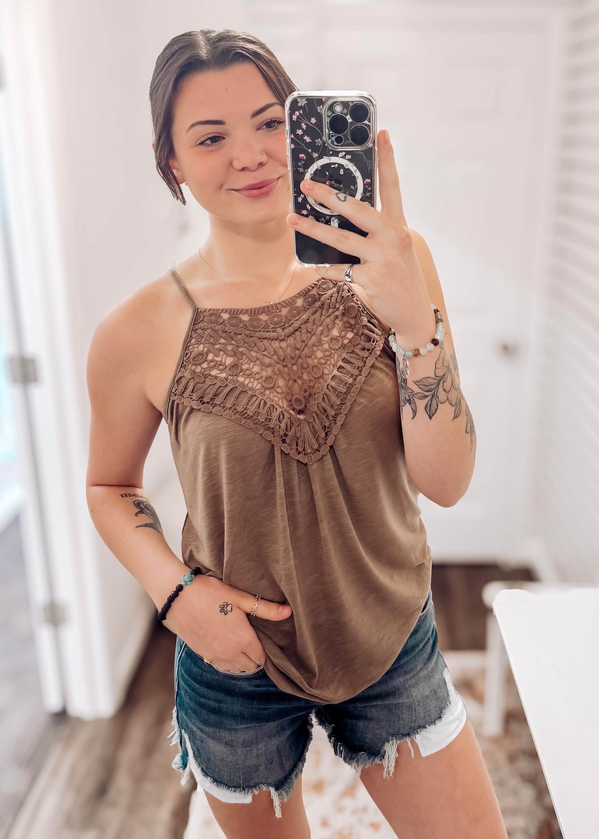 Woman taking a mirror selfie wearing a mocha brown halter tank top with intricate crochet lace detailing across the neckline and chest. The lightweight, flowy fabric drapes softly with a relaxed fit. Thin spaghetti straps and subtle pleating add to its breezy, feminine vibe. She pairs it with high-rise, frayed hem denim shorts—perfect for a casual yet stylish summer outfit.