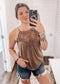Woman taking a mirror selfie wearing a mocha brown halter tank top with intricate crochet lace detailing across the neckline and chest. The lightweight, flowy fabric drapes softly with a relaxed fit. Thin spaghetti straps and subtle pleating add to its breezy, feminine vibe. She pairs it with high-rise, frayed hem denim shorts—perfect for a casual yet stylish summer outfit.