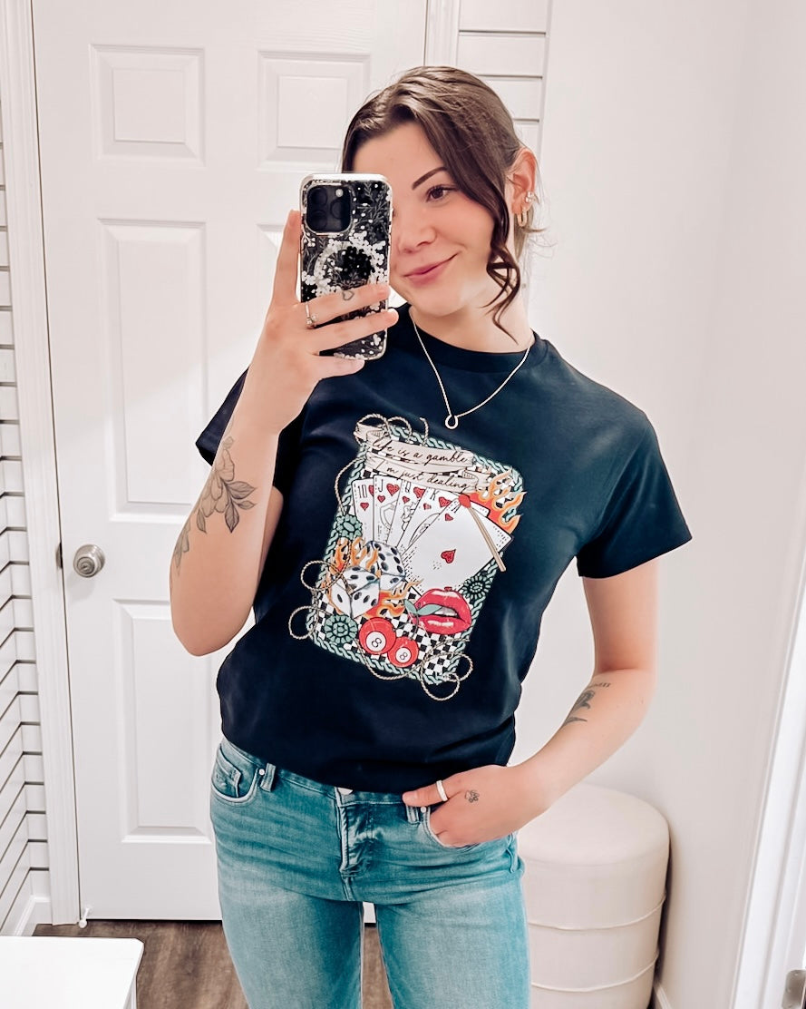Black graphic tee featuring a bold vintage tattoo-inspired design with playing cards, flames, dice, pool balls, checkerboard accents, and red lips. Banner at the top reads “Life is a gamble, I’m just dealing.” Perfect for adding a little edge and attitude to your casual wardrobe—ideal for concerts, game nights, or days when you’re feeling a little wild.