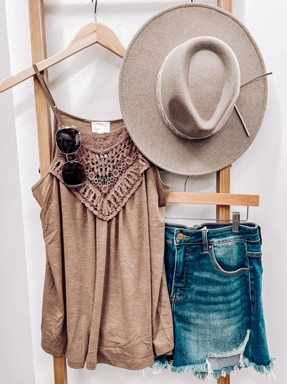 Woman taking a mirror selfie wearing a mocha brown halter tank top with intricate crochet lace detailing across the neckline and chest. The lightweight, flowy fabric drapes softly with a relaxed fit. Thin spaghetti straps and subtle pleating add to its breezy, feminine vibe. She pairs it with high-rise, frayed hem denim shorts—perfect for a casual yet stylish summer outfit.