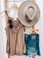 Woman taking a mirror selfie wearing a mocha brown halter tank top with intricate crochet lace detailing across the neckline and chest. The lightweight, flowy fabric drapes softly with a relaxed fit. Thin spaghetti straps and subtle pleating add to its breezy, feminine vibe. She pairs it with high-rise, frayed hem denim shorts—perfect for a casual yet stylish summer outfit.