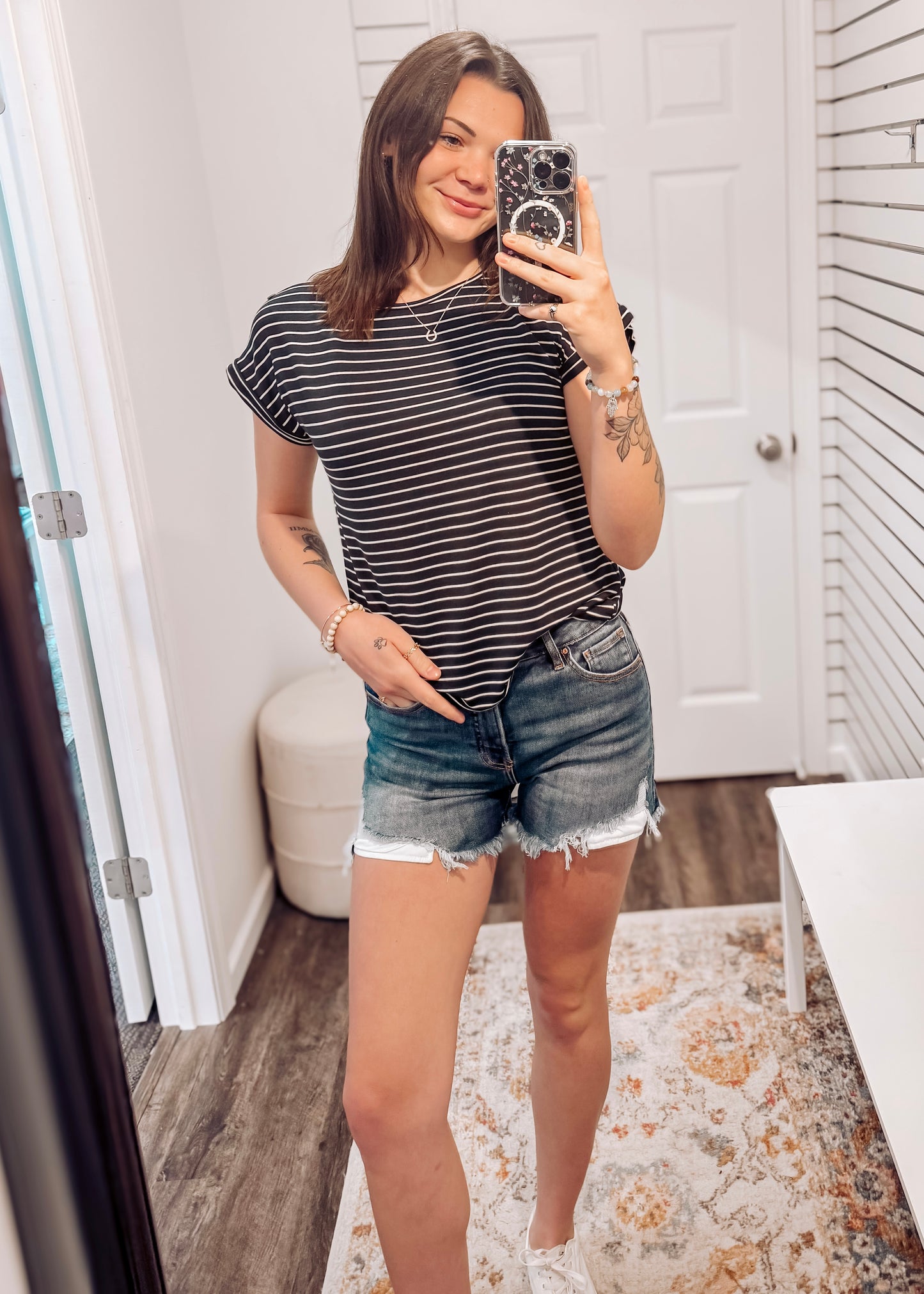 Woman wearing a black short-sleeve top with thin white horizontal stripes, a classic crew neckline, and rolled cuff sleeves with button accents. The top features a relaxed, drapey fit that pairs effortlessly with white cuffed shorts for a crisp, casual look. Perfect for everyday wear, this lightweight striped tee brings timeless style and comfort to your summer wardrobe.