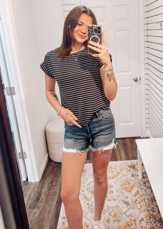 Woman wearing a black short-sleeve top with thin white horizontal stripes, a classic crew neckline, and rolled cuff sleeves with button accents. The top features a relaxed, drapey fit that pairs effortlessly with white cuffed shorts for a crisp, casual look. Perfect for everyday wear, this lightweight striped tee brings timeless style and comfort to your summer wardrobe.