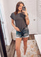 Woman wearing a black short-sleeve top with thin white horizontal stripes, a classic crew neckline, and rolled cuff sleeves with button accents. The top features a relaxed, drapey fit that pairs effortlessly with white cuffed shorts for a crisp, casual look. Perfect for everyday wear, this lightweight striped tee brings timeless style and comfort to your summer wardrobe.