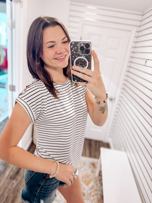 Woman wearing a white short-sleeve top with thin black horizontal stripes and rolled cuffs. The shirt features a relaxed, flowy fit with a crew neckline and button detail on the sleeves. Styled with white cuffed shorts, this effortless everyday tee is perfect for warm weather, offering a classic nautical-inspired look that’s both casual and polished.