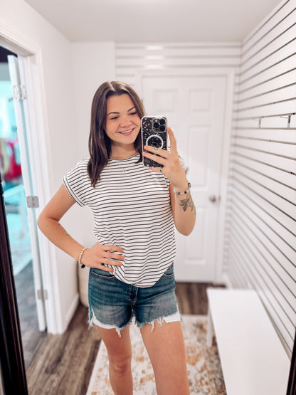 Woman wearing a white short-sleeve top with thin black horizontal stripes and rolled cuffs. The shirt features a relaxed, flowy fit with a crew neckline and button detail on the sleeves. Styled with white cuffed shorts, this effortless everyday tee is perfect for warm weather, offering a classic nautical-inspired look that’s both casual and polished.
