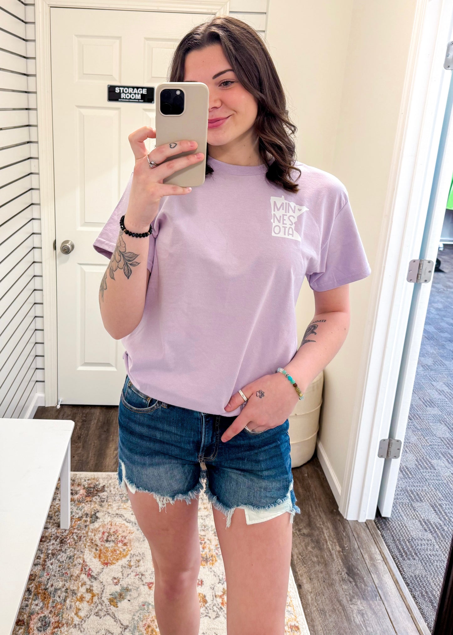 Woman wearing a relaxed-fit lavender graphic tee with a minimalist white design on the left chest that reads “MINNESOTA” stacked vertically within the outline of the state. Styled casually with frayed denim shorts and natural waves, this laid-back look is perfect for repping state pride with a soft pop of pastel color. Ideal for everyday errands, casual lake days, or chill weekends in the Midwest.